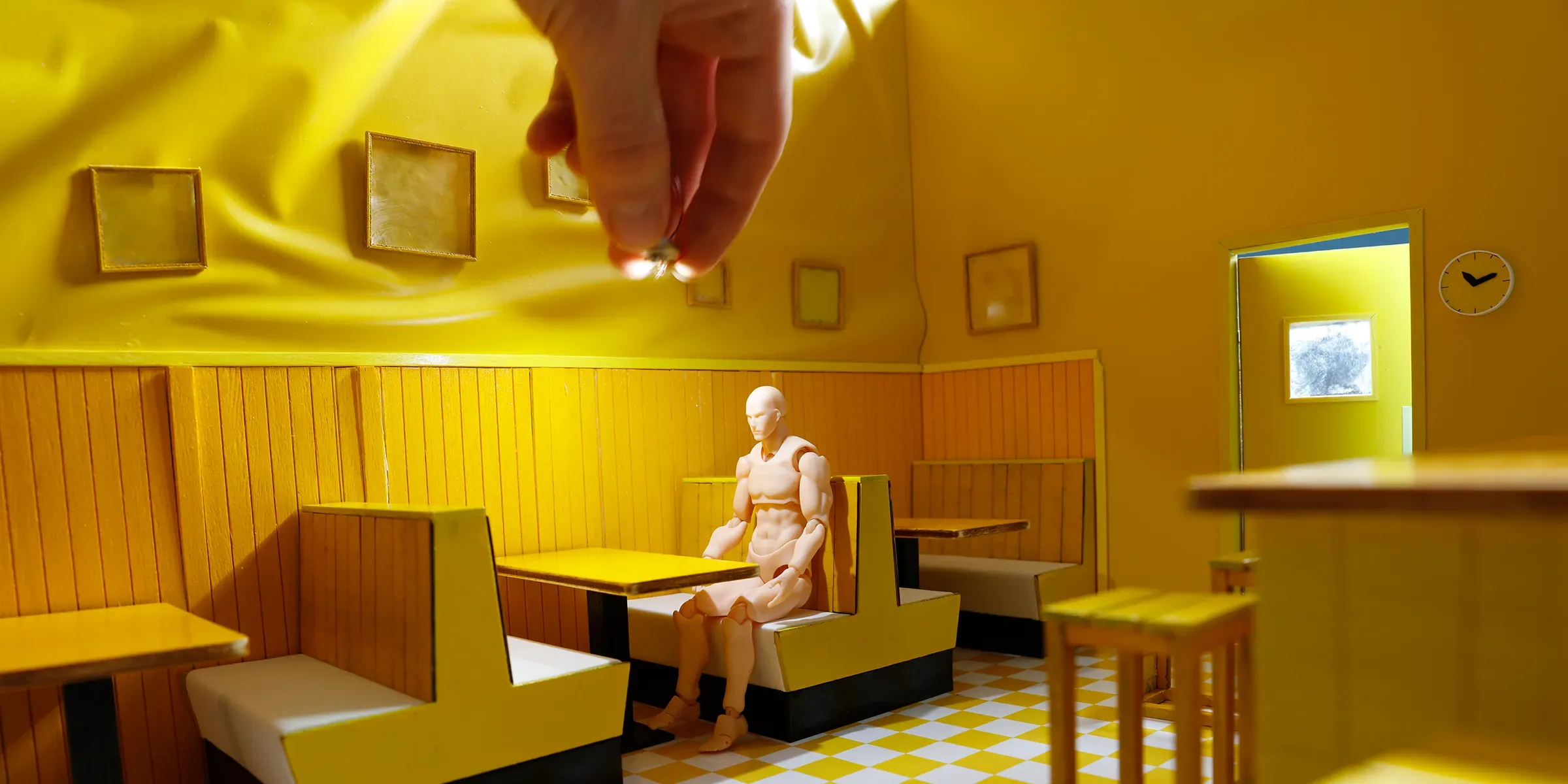A hand adjusts a lamp above a figure in a yellow miniature diner set